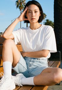 Jeune femme en t-shirt blanc, short en denim et baskets blanches assise sur un banc en bois en plein air avec des palmiers en arrière-plan.