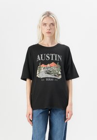 Juodas per didelis marškinėliai su grafiniu spausdinimu, vaizduojančiu kelią, kalnus ir kaktusus, su tekstu "AUSTIN TEXAS" kontrastingomis spalvomis.
