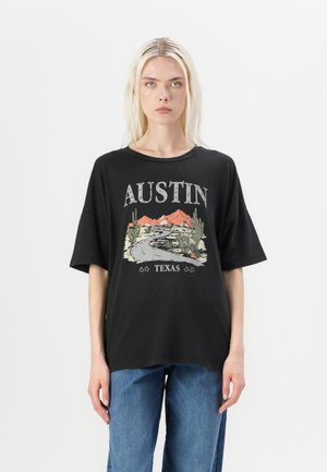 T-shirt noir surdimensionné avec un imprimé graphique représentant une route, des montagnes et des cactus, avec le texte "AUSTIN TEXAS" dans des couleurs contrastées.
