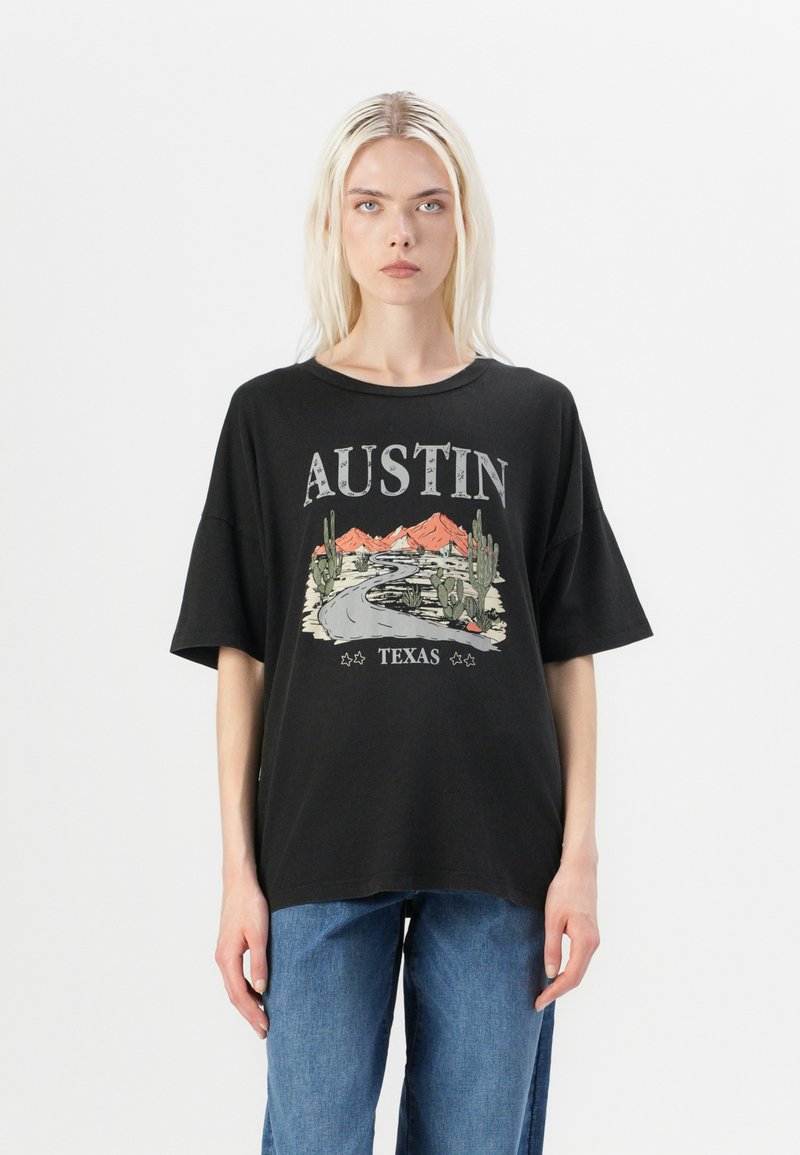 Juodas per didelis marškinėliai su grafiniu spausdinimu, vaizduojančiu kelią, kalnus ir kaktusus, su tekstu "AUSTIN TEXAS" kontrastingomis spalvomis.