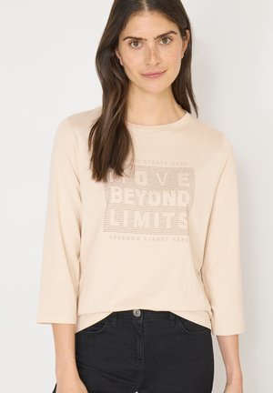 Chemise manches longues beige clair avec un col rond, présentant un imprimé graphique texturé dans des nuances de gris, associée à un pantalon noir.