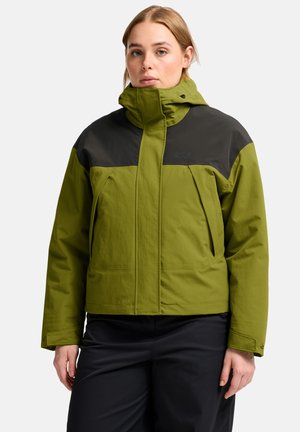 Vrouw draagt een tweekleurige olijfgroene en zwarte waterdichte jas met hoge kraag en zwarte broek, staand tegen een effen achtergrond.