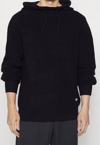 Hoodie noir en tricot avec un motif texturé, cordon de serrage et poignets côtelés. Dispose d'un patch logo à l'ourlet. Silhouette droite.