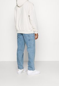 Felpa con cappuccio beige chiaro con coulisse e polsini a coste, abbinata a jeans larghi azzurri con tasche cargo e sneakers bianche.