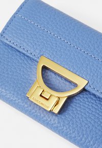 Cartera de cuero texturizado azul claro con un cierre metálico dorado que presenta un diseño geométrico y el nombre de la marca "Coccinelle" grabado.