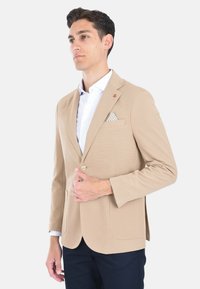 Blazer beige con una finitura testurizzata, un bottone singolo, due tasche frontali e un fazzoletto da taschino a contrasto con un motivo a pois.