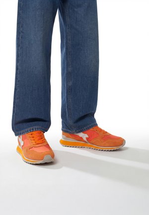 Persoon draagt wijde blauwe jeans en feloranje sneakers met wit sterdetail, staand op een witte vloer.