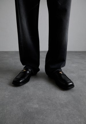Black leather loafers with a gold emblem, featuring a smooth texture and square toe, worn with dark denim pants.