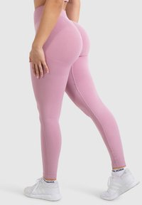 Leggings rosa a vita alta realizzati in tessuto elasticizzato, con una texture liscia, un design supportivo e scarpe da ginnastica bianche a contrasto.