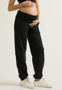 Zwarte zwangerschaps sweatpants met een brede elastische tailleband, gecuffte enkels en zijzakken, gecombineerd met witte sneakers.