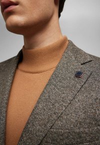 Brauner gestrickter Rollkragenpullover unter einem strukturierten grauen Blazer mit einem kleinen schwarzen und roten Revers-Pin, der eine Nahaufnahme der Stoffe zeigt.