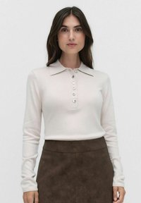 Vaalean beige pitkähihaisessa polo-neuleessa on kaulus ja kuusi nappia, yhdistettynä tummanruskeaan tekstuuriin midi-hameeseen. Kangas on sileä.
