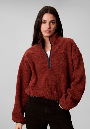 Frau trägt einen rostfarbenen Fleece-Pullover mit schwarzem Halb- Reißverschluss und schwarze Hose, steht vor schlichtem grauem Hintergrund.