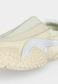 Gros plan d'une chaussure beige et blanche sans lacets avec une semelle en caoutchouc texturée et un motif de vague cousu sur le côté.