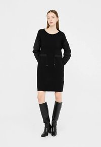 Melna adīta kleita ar apaļu izgriezumu kaklā, garām piedurknēm un divām priekšējām kabatām. Valkāta kopā ar melniem zābakiem līdz ceļiem. Gluda tekstūra, vienkāršs dizains.