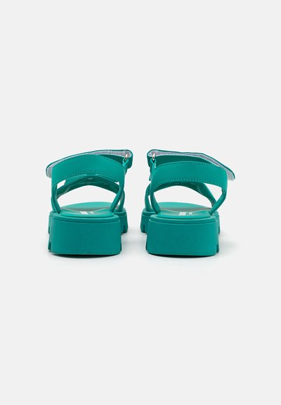 Tealfärgade sandaler med en kraftig sula, utrustade med dubbla justerbara remmar och en texturerad fotbädd. Bakifrån visas rundade klackar och öppen design.