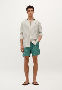 Chemise à manches longues rayée dans des couleurs claires, associée à un short de bain vert avec des motifs de têtes de mort et os croisés jaunes. Tongs noires.