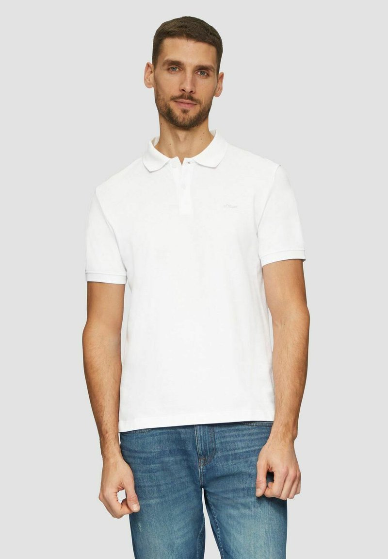 Weißes Poloshirt aus Baumwolle mit klassischem Kragen, kurzen Ärmeln und dezentem Markenlogo auf der linken Brust. Kombiniert mit blauen Jeans.