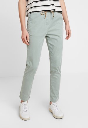 Persoon die lichtgroene broek met trekkoord, witte sneakers en een zwart-wit gestreept shirt draagt, staand op een effen witte vloer.