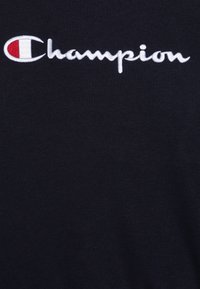 Tumši zils T-krekls ar izteiktu balto izšūto "Champion" logo, kas papildināts ar sarkanu un baltu apļa akcentu. Mīksta kokvilnas tekstūra.