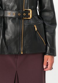 Black leather jacket with a fitted design, gold hardware, front zip closure, and side pockets. The jacket features a belted waist for shape.