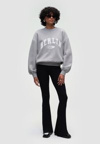 Szara bluza z napisem "BERLIN", w luźnym kroju, z prążkowanymi mankietami, w zestawieniu z czarnymi prążkowanymi legginsami w kształcie dzwonów oraz czarnymi butami.
