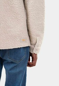 Beige fleece-takki, jossa on teksturoitu pinta ja tarralipuke, jossa on kultainen logo, yhdistetty tummansinisiin farkkuihin, ja käsi lepää reidellä.
