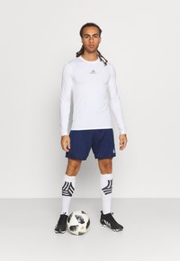 Weißes Langarmhemd mit grauem Logo, marineblaue Shorts, schwarze Fußballschuhe und gemusterte kniehohe Socken. Hält einen schwarz-weißen Fußball.