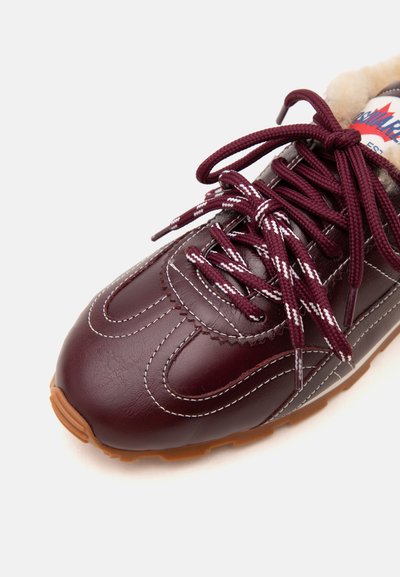Gros plan sur une basket en cuir bordeaux avec des coutures blanches, des lacets bordeaux à motifs et une semelle en caoutchouc beige, montrant partiellement un intérieur doublé de fourrure.