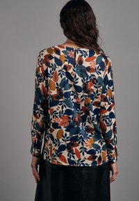 Blouse à manches longues sur une base crème avec un motif de feuilles colorées en bleu, orange et vert. Elle présente un col rond et une coupe décontractée.