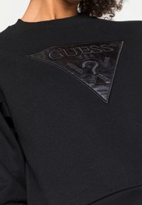 Sudadera negra con un parche de cuero en relieve en forma de triángulo que lleva el texto "GUESS" y un diseño de signo de interrogación. Tejido texturizado, puños acanalados.