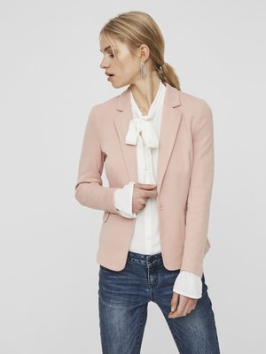 Woman wearing light pink blazer, white blouse with bow tie, blue jeans, and statement earrings, looking to the side against a plain background.
