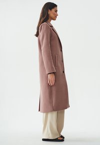 Lungo cappotto su misura in morbido tessuto rosa antico. Presenta un colletto a punte, quattro bottoni neri e tasche laterali. Abbinato a pantaloni beige chiaro.