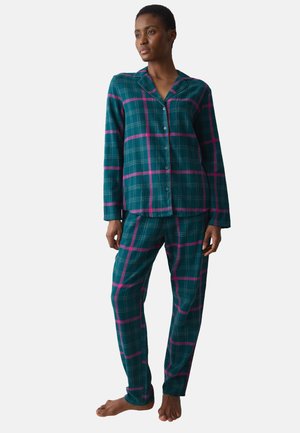 Personne debout pieds nus portant un haut à manches longues boutonné à carreaux bleu sarcelle et rose avec un pantalon assorti sur fond blanc.