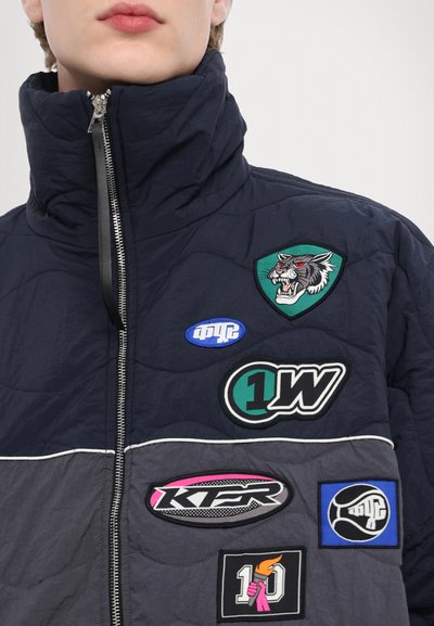 Quilted navy jacket with high collar, featuring six embroidered patches in various colors and shapes, including a wolf and logos. Zippered front.