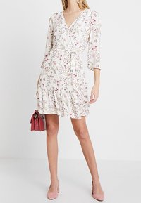 Floral wrap dress in light fabric featuring a V-neck, three-quarter sleeves, and ruffled hem. Accompanied by a pink bag and heels.