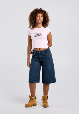 Vrouw staat met het gezicht naar voren, draagt een roze cropped t-shirt met cassetteprint, knielange spijkershort en beige veterschoenen.