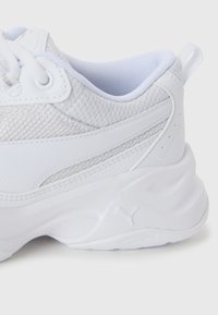 Baskets blanches de sport avec dessus en mesh et cuir, semelle épaisse, design à lacets, et languette en tissu à l'arrière.