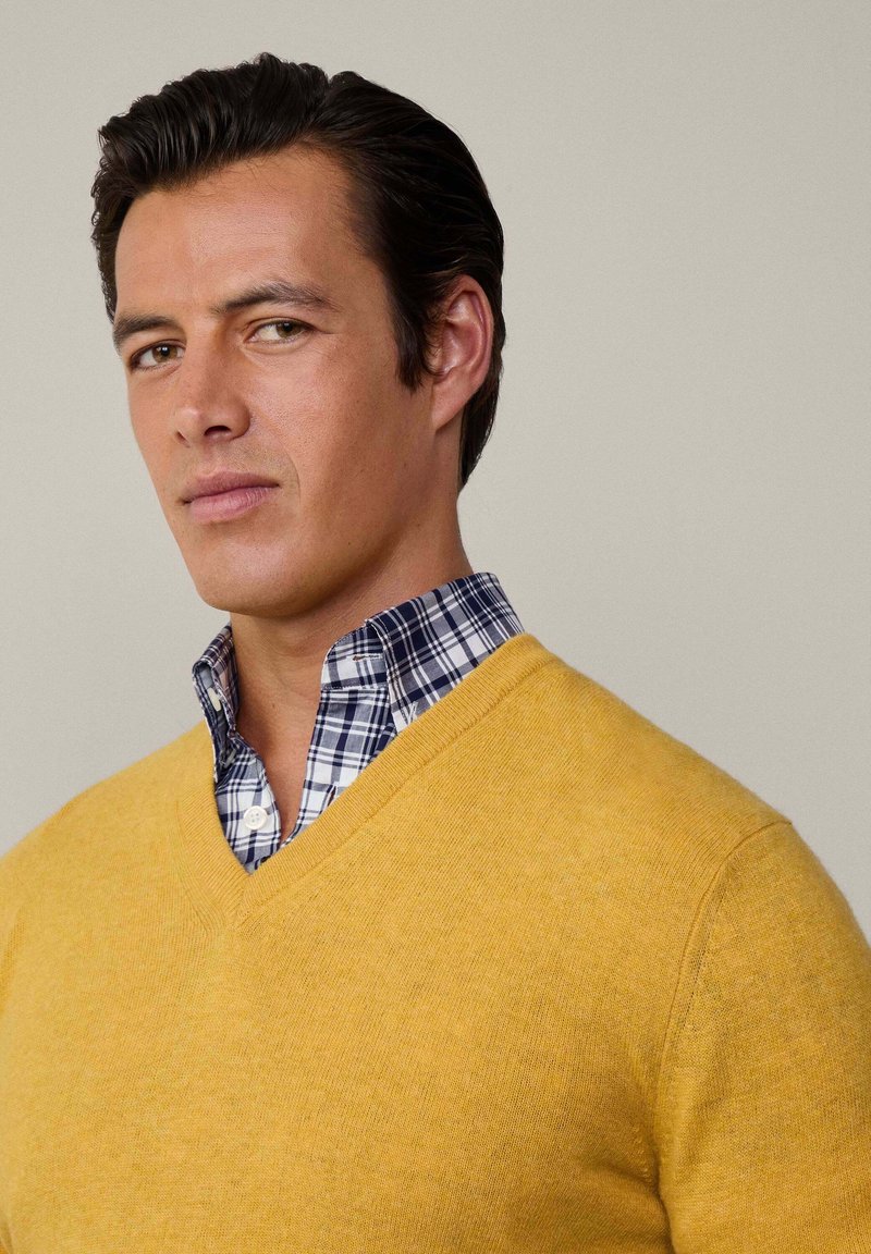 Pull en V jaune en matériau doux superposé sur une chemise à carreaux bleue et blanche. Texture lisse, design ajusté et couleurs contrastantes.