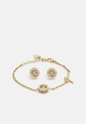 Armband van goudkleurig metaal van Guess en bijpassende oorstekers, elk met heldere kristalcentra en gegraveerde merknaamaccenten.