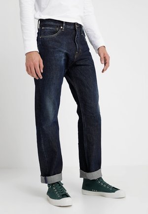 Man draagt donkere opgerolde jeans, witte lange mouwen shirt en groene sneakers met witte zolen, staand tegen een effen achtergrond.