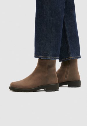 Brown ankle boots with side zipper worn with dark blue denim wide-leg jeans on light gray background.