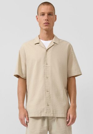 Man staat in een beige overhemd met korte mouwen en knopen en bijpassende korte broek over een wit ondershirt tegen een effen lichte achtergrond.