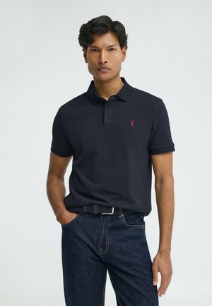 Man draagt een donkerblauw poloshirt met een klein rood logo, ingestopt in donkere spijkerbroek met een zwarte gevlochten riem, staand tegen een witte achtergrond.