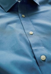 Modra srajca s kratkimi rokavi in gladko teksturo, ki ima klasičen ovratnik, bele gumbe ter kontrastno belo šivanje ob zapenjanju.