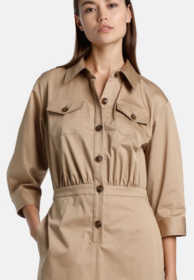 Tan utility dress with a collared neckline, front button closure, two chest pockets, and elastic waist for a fitted silhouette.