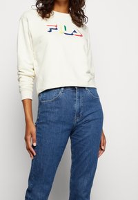 Kvinna som bär en krämfärgad FILA sweatshirt med flerfärgad logga och blå högmidjade jeans mot en helt vit bakgrund.