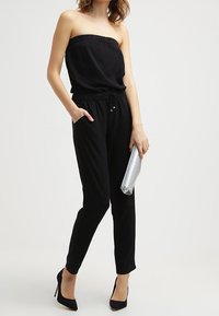 Combinaison noire sans bretelles avec un col élastique, des jambes cintrées et une taille à cordon. Associée à une pochette argentée et des talons noirs.