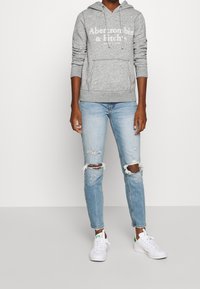 Felpa grigia con cappuccio e tasca frontale, con testo bianco. Indossata con jeans azzurri chiari strappati e sneakers bianche con accenti verdi.