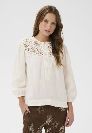 Frau mit langen braunen Haaren, die eine weiße Bluse mit Spitzenkragen und braune Hose mit Seestern- und Korallenmustern trägt, posiert vor einem einfarbigen Hintergrund.